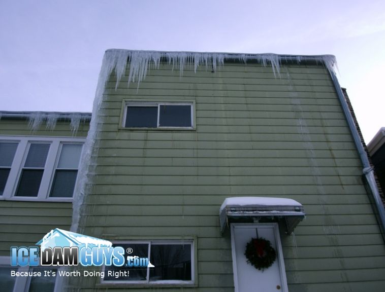The Ice Dam Guys help apartment building managers and owners with ice dams. In this photo we see a green multi-family home that has icicles coming from an ice dam hanging over the gutters. There's a wreath on the door, so it must be the holidays.