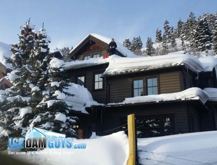 This photo shows what appears to be a large mountain lodge or home with snow on the roof. There is a man up there with a shovel, shoveling snow off the roof to expose an ice dam below. He is preparing for ice dam removal in Ontario, Canada.