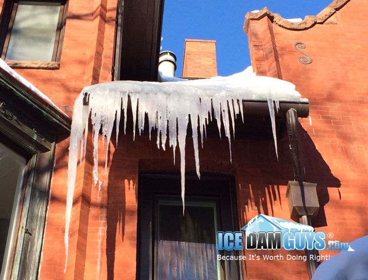 The photo is of a historic home, made of brick and stone, that has an ice dam hanging over the front stoop. The home is ornately designed and the ice dam is hanging several feet at it largest.