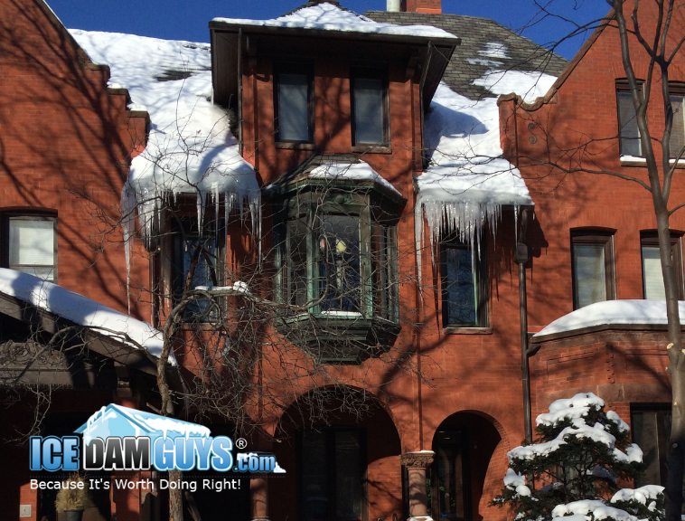 This is an older home, made of red bricks, that has ornate architecture and looks to be historic. It has many icicles from an ice dam hanging off its steep roof. The Ice Dam Guys' logo is superimposed over the image.