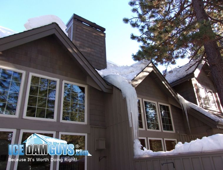 The Ice Dam Guys regularly have to help vacation rental owners address their ice dams. Like the photo here, many rentals are in the forest and nature. This photo has snow on a brown vacation property with icicles hanging down. There are evergreen trees around it.